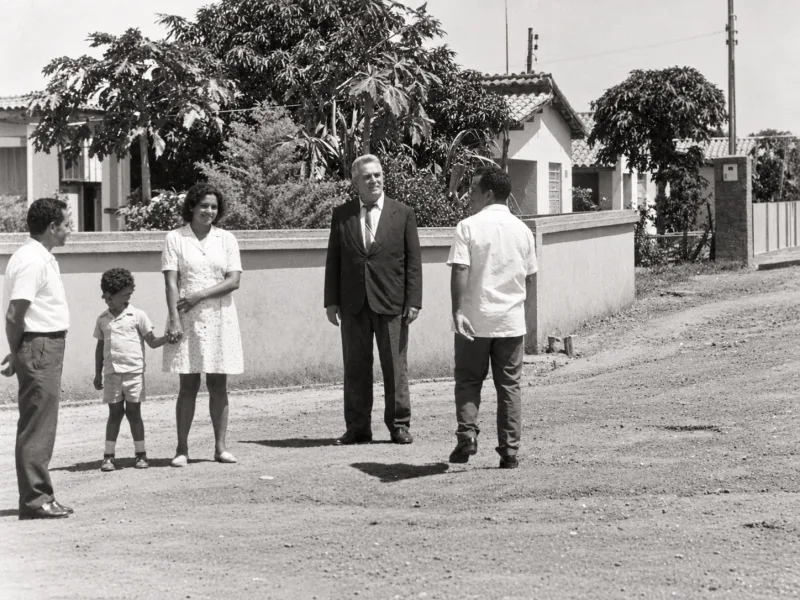 Zé Paulista de terno em rua de terra, ao lado de moradores, diante de casas baixas.