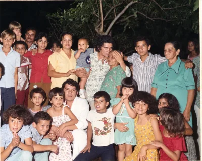 Adultos e crianças reunidos à noite sob as árvores, em foto colorida.