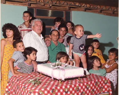 Família reunida ao redor de um bolo de aniversário, sobre mesa de toalha xadrez.