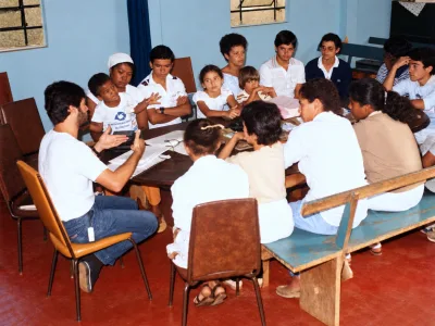 Pessoas sentadas em bancos e cadeiras durante uma reunião no salão da casa.
