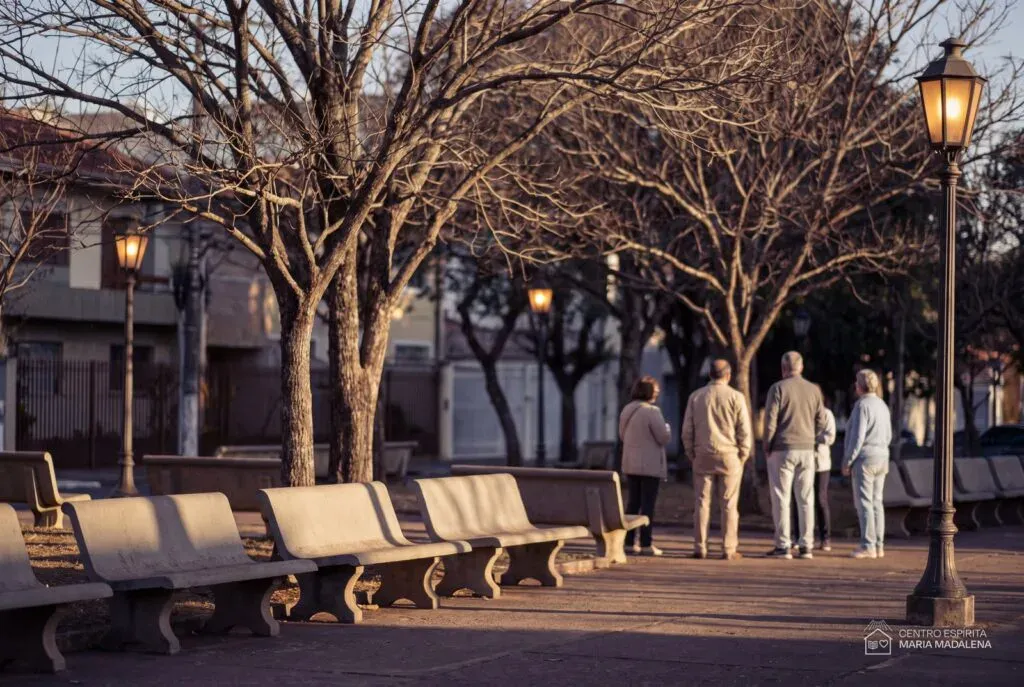 Praça arborizada de bairro ao entardecer, com bancos vazios e pessoas de costas conversando ao fundo
