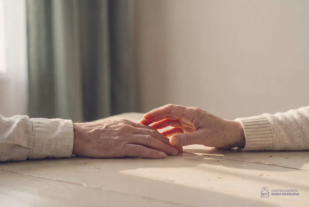 Duas mãos de pessoas idosas sobre uma mesa de madeira clara, quase se tocando, na luz de uma janela