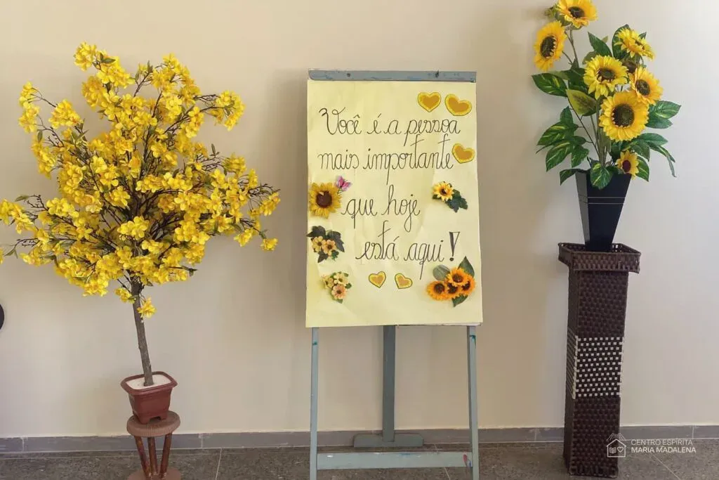 Cartaz feito à mão no CEMA com a frase "Você é a pessoa mais importante que hoje está aqui", decorado com girassóis e corações, entre arranjos de flores amarelas