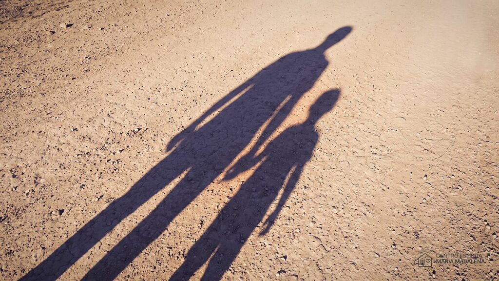 Sombras de um adulto e de uma criança caminhando lado a lado num caminho de terra ao fim da tarde