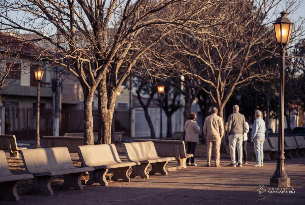 Praça arborizada de bairro ao entardecer, com bancos vazios e pessoas de costas conversando ao fundo