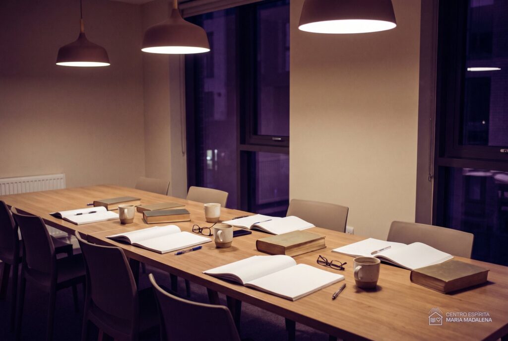 Mesa comprida de estudo à noite com livros abertos, cadernos e xícaras, cadeiras afastadas