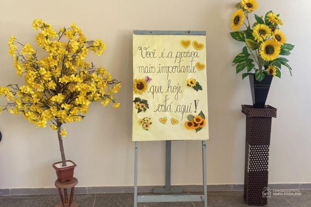 Cartaz feito à mão no CEMA com a frase "Você é a pessoa mais importante que hoje está aqui", decorado com girassóis e corações, entre arranjos de flores amarelas